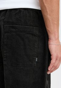 Los pantalones de pana negros cuentan con rayas verticales, un bolsillo trasero y una etiqueta en el lado. La tela parece texturada y suave.