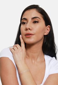 Zilveren bracelet met twee slanke banden, gedragen om een pols, die een witte shirt met een scherpe pasvorm aanvult tegen een effen achtergrond.