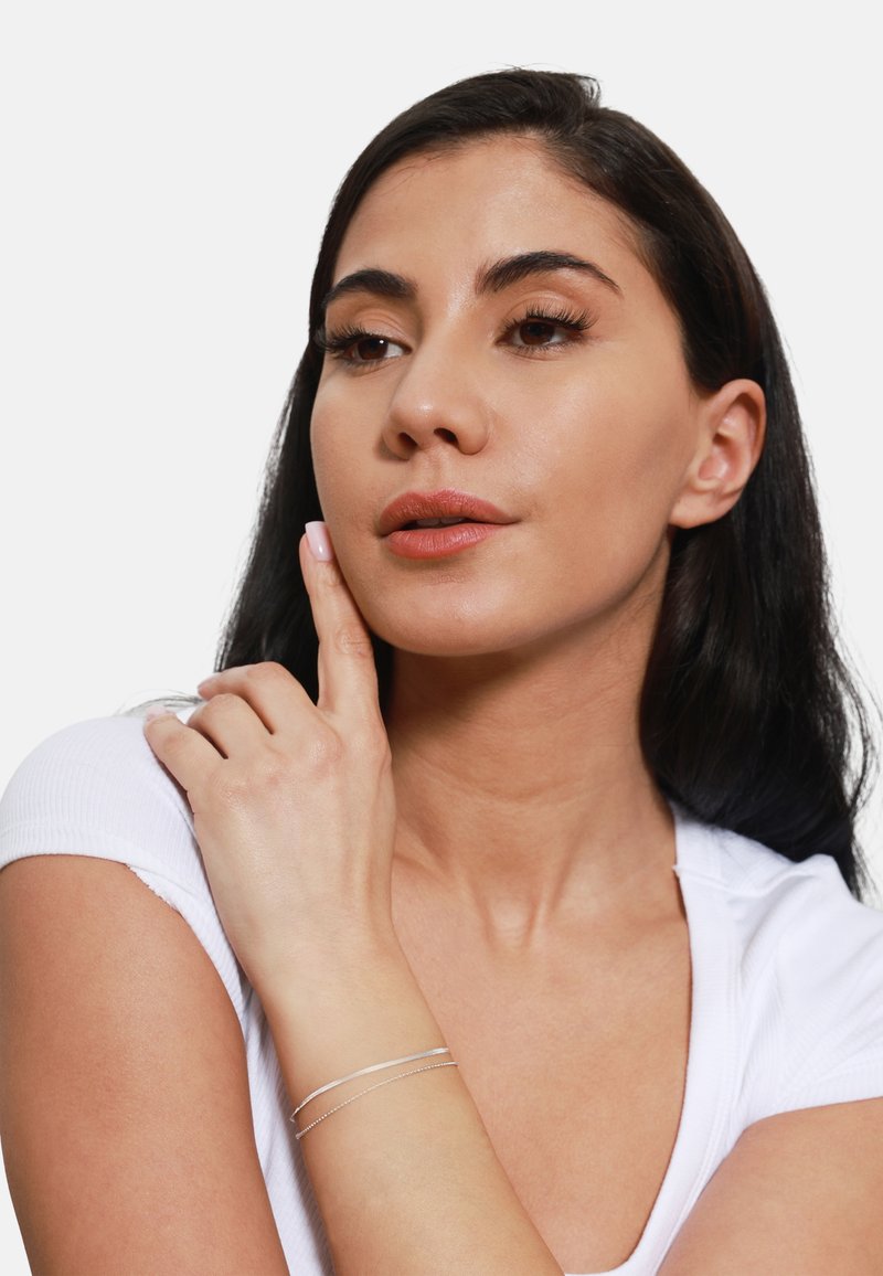 Zilveren bracelet met twee slanke banden, gedragen om een pols, die een witte shirt met een scherpe pasvorm aanvult tegen een effen achtergrond.
