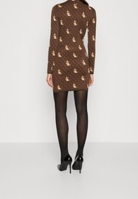 Robe marron à manches longues avec un motif répétitif de dessins crème et rouges, coupe ajustée, portée avec des collants noirs transparents et des talons hauts.