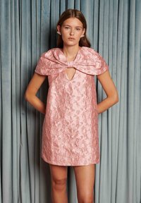 Kort rosa kjole med stor sløyfe på kragen, intrikat blomsterbroderi og V-hals. Glatt tekstur, lengde over kneet.