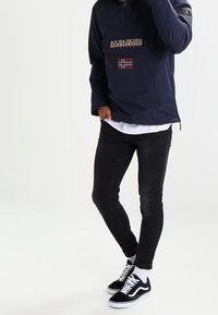 Chaqueta de pullover de color navy con media cremallera y bolsillo frontal, que presenta un parche con el logo y la bandera noruega; combinada con vaqueros ajustados negros y zapatillas Vans negras.