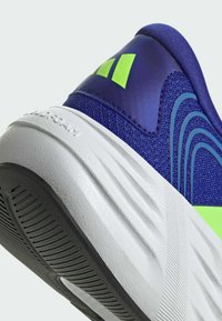 Gros plan du talon d'une chaussure de sport bleue avec un logo vert fluo, une semelle intermédiaire blanche texturée et une semelle extérieure en caoutchouc noir avec des motifs antidérapants.