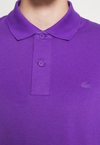 Camisa polo morada con una tela texturizada, que presenta un cierre de tres botones y un logo en relieve en el lado izquierdo del pecho.