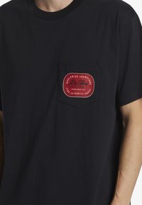 Svart bomull t-shirt med en röd cirkulär patch med vit text på vänster bröstficka. Kortärmad och design med rund hals.