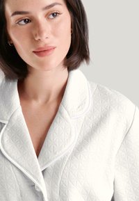 Chaqueta blanca texturizada con patrones de diamante, cuello chal y ribete contrastante. Vista en primer plano, destacando los detalles de la tela y los botones.