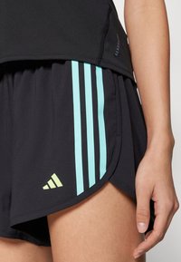 Los pantalones cortos deportivos negros presentan rayas verticales de color azul claro y un pequeño logo en amarillo-verde en el dobladillo. El diseño está ajustado con una tela que absorbe la humedad.