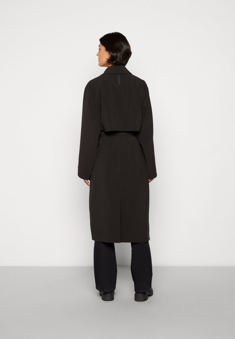 Lungo trench coat nero con vita cintata, colletto e spacco sul retro, realizzato in tessuto liscio. Indossato su pantaloni neri e stivali.