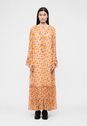 Žena s dlhými čiernymi vlasmi v broskyňových dlhých rukávoch maxi šatách s oranžovým kvetinovým vzorom a volánikovými detailmi, v kombinácii s čiernymi topánkami.