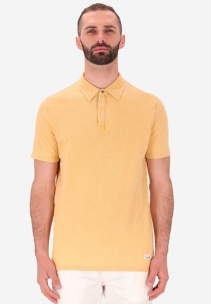 Polo giallo con una trama morbida, colletto con bottoni e maniche corte. Presenta un'etichetta piccola sull'orlo, indossato con pantaloni bianchi.
