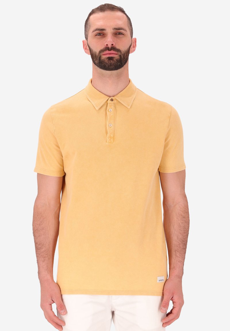 Polo giallo con una trama morbida, colletto con bottoni e maniche corte. Presenta un'etichetta piccola sull'orlo, indossato con pantaloni bianchi.