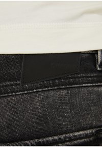 Vaqueros de mezclilla negra con una superficie texturizada y una etiqueta de parche con texto en relieve. La parte superior presenta un dobladillo de camiseta de color claro.