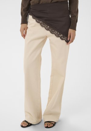 Persona che indossa pantaloni crema a gamba larga con sopra una gonna a portafoglio asimmetrica marrone scuro bordata di pizzo e sandali neri con cinturini.