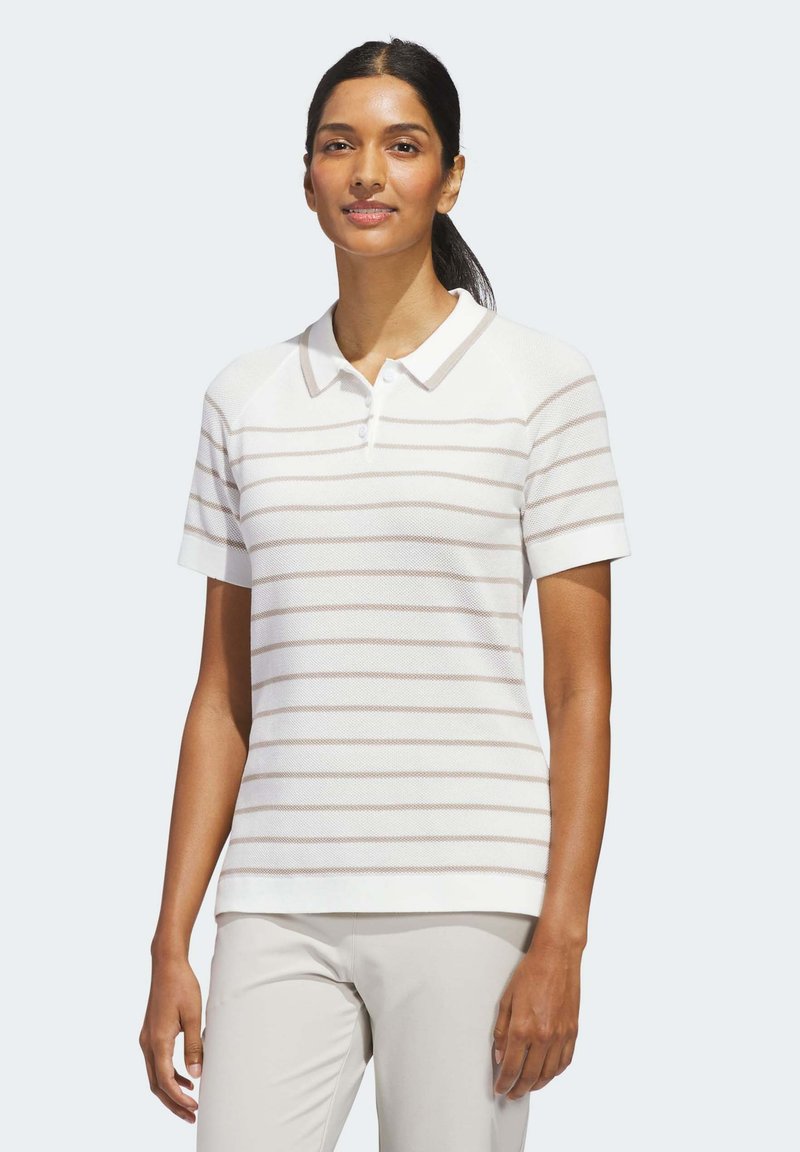 Γυναίκα με σκούρα μαλλιά, πιασμένα σε χαμηλή αλογοουρά, που φοράει λευκό polo μπλουζάκι με κοντά μανίκια και ανοιχτές οριζόντιες ρίγες, καθώς και ανοιχτόχρωμο παντελόνι.