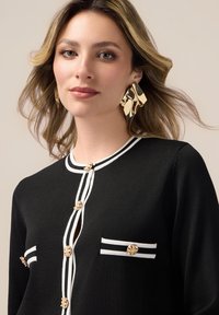 Cardigan nero con bordi bianchi, bottoni a forma di fiore dorati e una tasca sul petto. Modella con orecchini dorati texturizzati. Tessuto liscio e aderente.