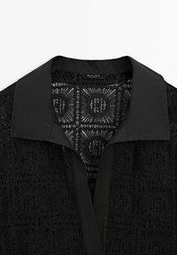 Chemise noire à col avec un motif complexe en dentelle ajourée formant des dessins géométriques carrés sur le tissu.