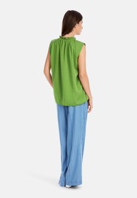 Grüne ärmellose Bluse mit gerafftem Ausschnitt; kombiniert mit weit geschnittenen hellblauen Hosen. Der Stoff wirkt glatt und leicht.