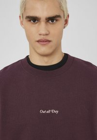 Jeune personne avec les cheveux blonds courts portant une chemise bordeaux avec le texte "Out all Day", fond clair uni.