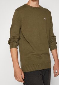 Oliivinvihreä neulepaita, jossa on resorit hihansuissa ja helmassa, ja rintakehässä pieni logopl�äntti. Yksinkertainen muotoilu ja teksturoitu kangas.