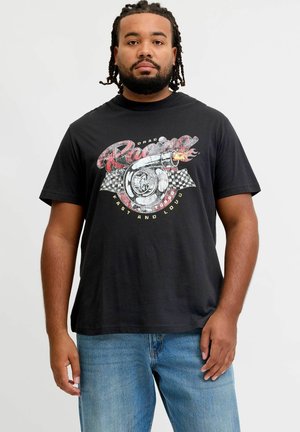 Mand med skæg og dreadlocks iført en sort t-shirt med en turbolader, ternede flag og teksten "Racing Fast and Loud", kombineret med blå jeans.