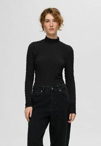 Juoda ilgomis rankovėmis palaidinė su tekstūruota, siuvinėta struktūra ir aukštu kaklu, derinama su juodomis aukštu liemeniu džinsais. Lygus audinio paviršius.