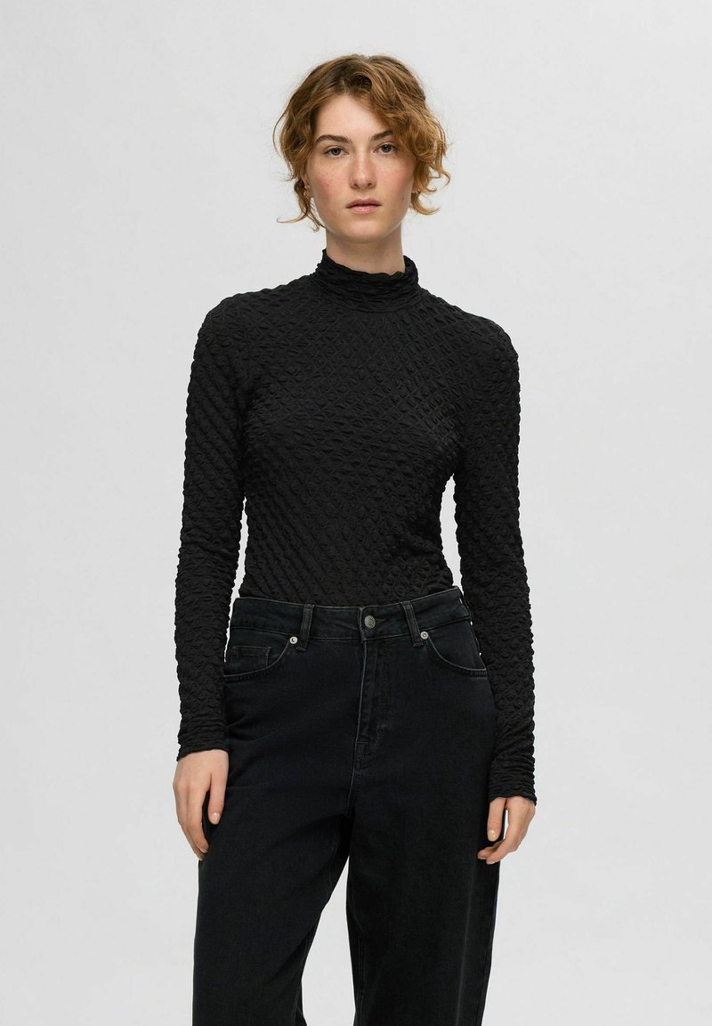 Juoda ilgomis rankovėmis palaidinė su tekstūruota, siuvinėta struktūra ir aukštu kaklu, derinama su juodomis aukštu liemeniu džinsais. Lygus audinio paviršius.