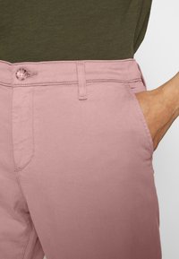 Ljusrosa bomullbyxor med knäppning, sidoficka och sömmar. Kombinerade med en mörkgrön top i tyg.