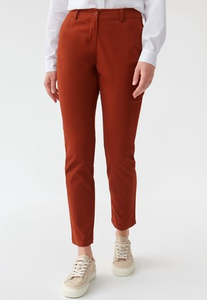 Pantaloni slim-fit color ruggine indossati con sneakers beige a fantasia e una camicia bianca a maniche lunghe, mostrati dalla vita ai piedi.