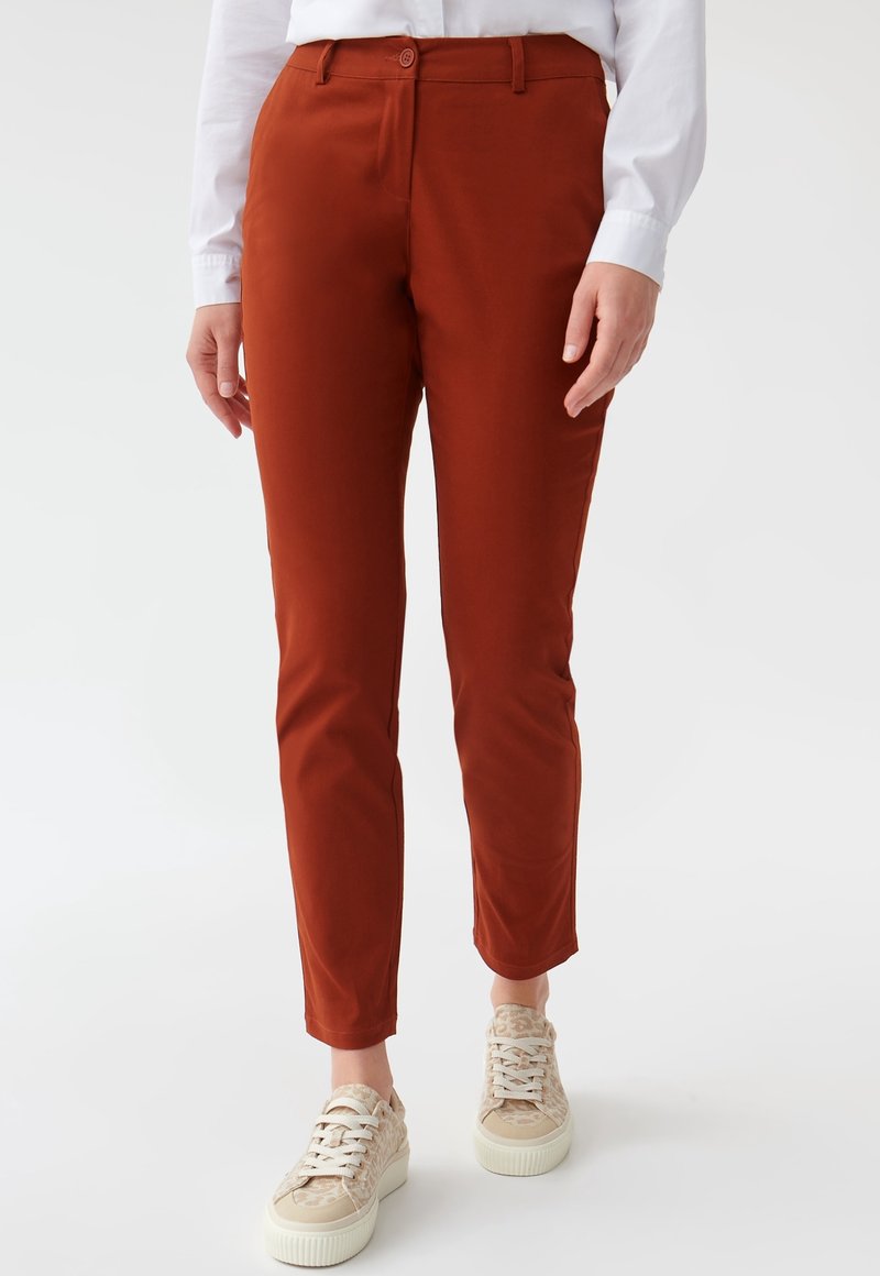Pantalon slim couleur rouille porté avec des baskets beige à motifs et une chemise blanche à manches longues, présenté de la taille aux pieds.