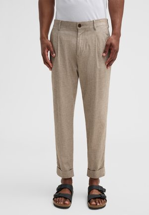 Beige Faltenhose aus einem strukturierten Stoff mit Knopfverschluss, seitlichen Taschen und umgeschlagenen Bündchen, kombiniert mit schwarzen Sandalen.
