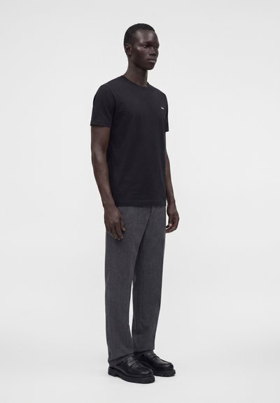 Homme debout portant un t-shirt noir à manches courtes, un pantalon gris et des mocassins noirs sur un fond gris clair uni.