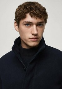 Marineblå kappa med hög krage, dragkedjestängning och en strukturerad svart tröja under. Modellen har en neutral ansiktsuttryck.