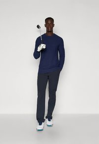 Μπλε σκούρο πουκάμισο με μακριά μανίκια, σκούρο παντελόνι, λευκά παπούτσια γκολφ με μπλε λεπτομέρειες· κρατώντας μπαστούνι γκολφ, φοράει λευκό γάντι.