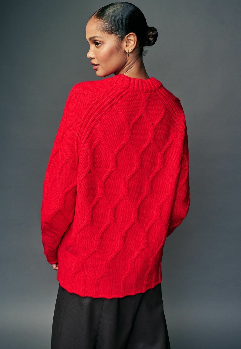 Maglione rosso lavorato a maglia con un motivo a diamante testurizzato, colletto e orlo a coste, vestibilità oversize, indossato sopra un indumento nero.