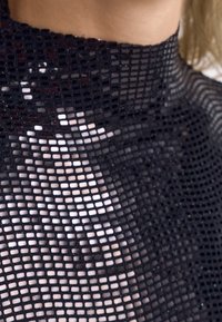 Primo piano di un tessuto a rete nero che copre la pelle, mostrando piccoli fori quadrati e parte del collo e della spalla di una donna.