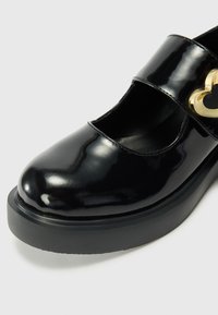 Melnās lakotas ādas Mary Jane kurpes ar apaļu purngali, bieza melna zole un zelta sirds formas sprādze uz siksniņas.