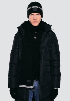 Muž nosící černou pletenou čepici, černý šál, černé rukavice, černou péřovou bundu a modré džíny na jednoduchém světlém pozadí.