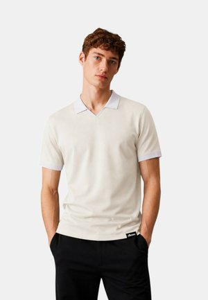 Giovane uomo che indossa una polo crema a maniche corte con collo bianco e pantaloni neri, in piedi con le mani in tasca davanti a uno sfondo semplice.