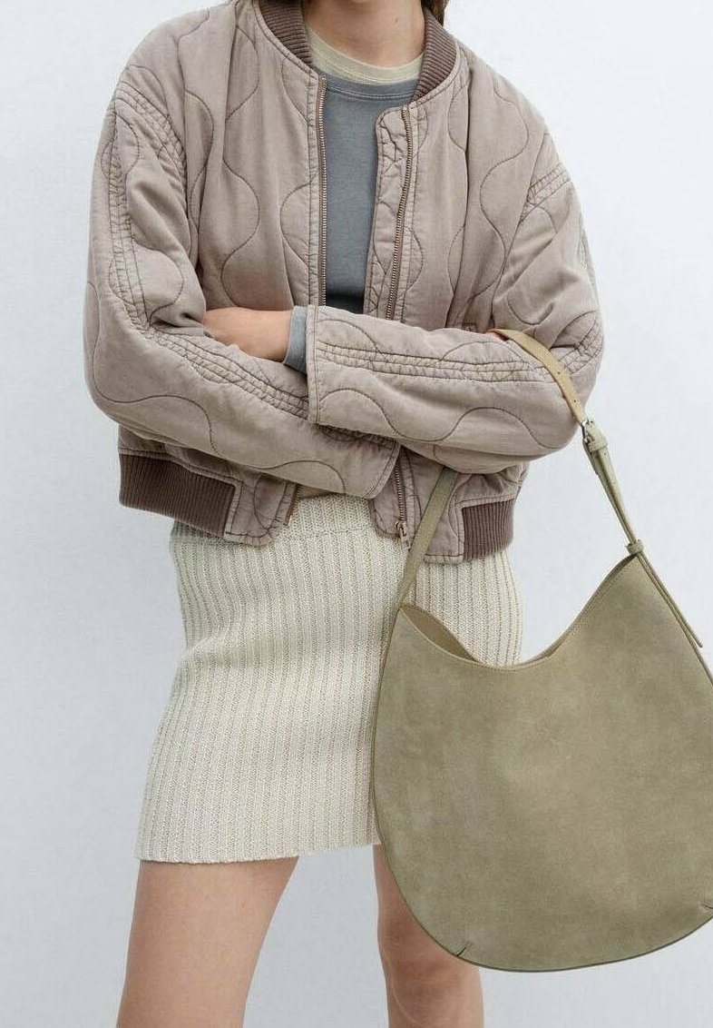 Personne portant une veste beige matelassée, un haut gris, une jupe en tricot crème, tenant un grand sac à bandoulière en daim vert olive avec une longue sangle.
