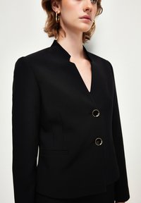 Czarna, dopasowana marynarka z dekoltem w kształcie litery V, okrągłym projektem bez kołnierza oraz dwoma okrągłymi złotymi guzikami. Posiada boczne kieszenie i gładką teksturę.
