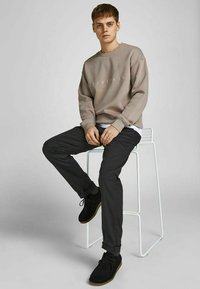 Lichtbruine sweatshirt met "ORIGINALS" gedrukt, zwarte slim-fit broek en zwarte suède schoenen, zittend op een witte kruk tegen een effen achtergrond.