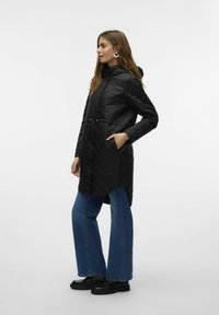 Zwarte gewatteerde jas met capuchon, cinched taille en zijzakken, gecombineerd met blauwe wijde jeans en zwarte platform schoenen.