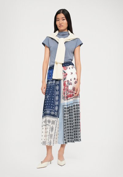 Donna in un top blu a maniche corte, una lunga gonna a motivo patchwork, un maglione bianco appoggiato sulle spalle e scarpe a punta bianche.