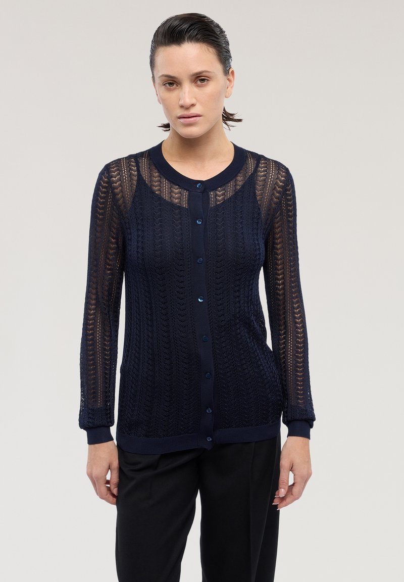 Mujer vistiendo un cárdigan de punto azul marino translúcido con mangas largas y botones, combinado con pantalones negros, de pie frente a un fondo liso.