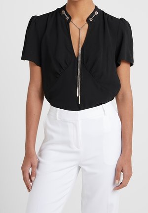 Vrouw draagt een zwarte blouse met korte mouwen en zilveren kettingdetail, gecombineerd met witte hoge taillebroeken, tegen een effen achtergrond.