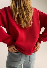 Czerwona bluza z luźnym krojem i ściągaczami, w połączeniu z jasnoniebieskimi dżinsami, z widocznymi detalami ściegów.