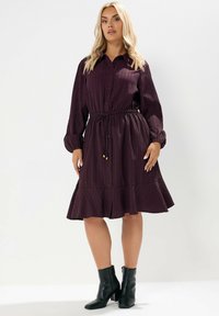 Vestito camicia viola scuro con texture a righe verticali, maniche lunghe, chiusura con bottoni e vita arricciata. Presenta un orlo svasato e viene indossato con stivaletti neri alla caviglia.