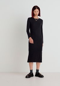 Black knitted long-sleeve dress with a fitted silhouette, knee-length hem, and subtle texture. Worn with chunky black shoes and gray socks.