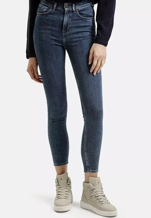 Kobieta w ciemnoniebieskich skinny jeansach i jasnobeżowych wysokich trampkach, stojąca na tle gładkiej białej ściany.