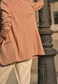 Różowy żakiet w odcieniu blush o fakturowanej tkaninie i luźnym kroju, z pojedynczą kieszenią, zestawiony z beżowymi spodniami, na tle kamiennej ściany.